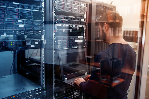 man works on laptop in front of server rack in data center